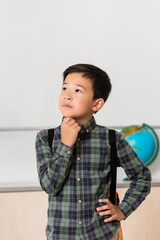 Thoughtful asian schoolboy with hand on hip in classroom