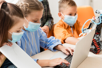 Selective focus of kids in medical masks using laptop and digital tablet near robot in school