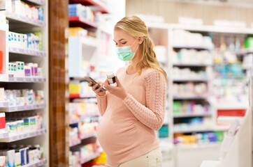 pregnancy, medicine and health concept - pregnant woman wearing protective medical mask for protection from virus disease with smartphone and drug at pharmacy