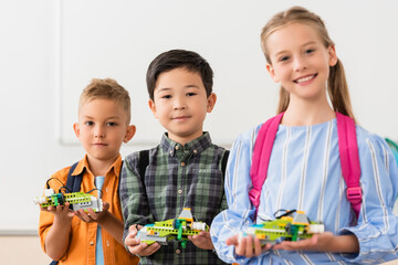 Selective focus of multiethnic pupils holding robots and looking at camera in stem school