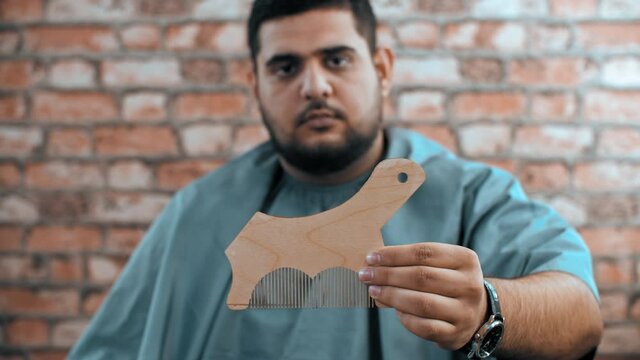 Serious bearded man holding beard shaping template. Close-up view of man in mantle sitting in barber shop and showing wooden beard shaping stencil