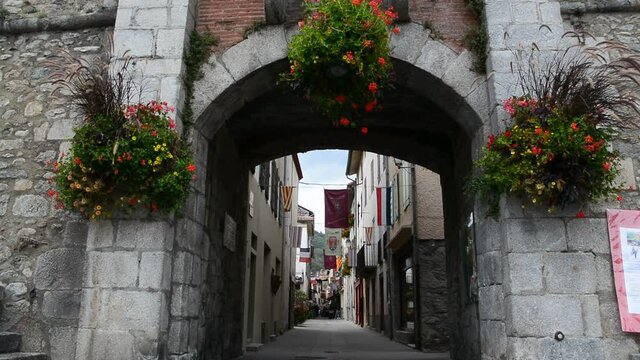 Still image of the main entrance of the Prats de Mollo la Preste fortress. From that angle, you can see red flowers that surround the arcade and the medieval street with flags of Catalonia and France