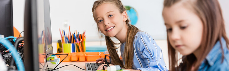 Fototapeta Panoramic shot of schoolgirl looking at camera near friend and computers in stem school obraz