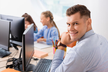 Obraz premium Selective focus of teacher looking at camera near computers and pupils in classroom