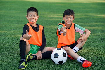 Joyful teenage male soccer players show ok sign resting on artificial turf of football field after...