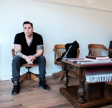 A tattooed man sits in a wooden chair looking our the window.