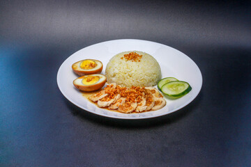 Hainanese chicken rice or steamed chicken rice with sweet soy sauce egg plus cucumber slices. Served on a white plate.
