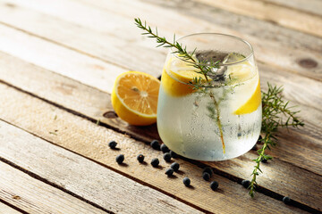Alcohol drink (gin tonic cocktail) with lemon, juniper branch,  and ice on rustic wooden table.