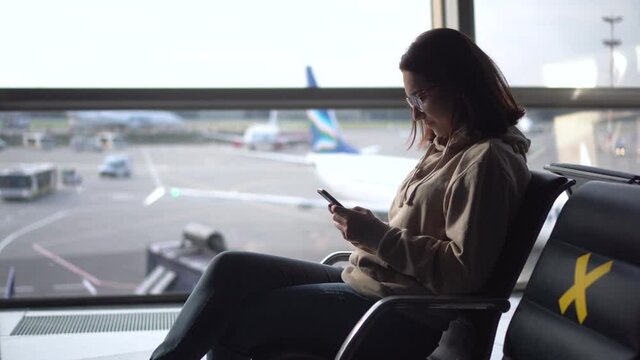 A young woman sits with a phone in her hands against the background of a window at the airport. The chairs are marked with crosses to maintain social distance.