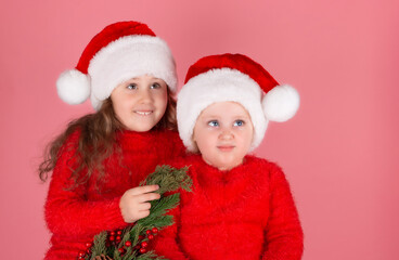 Two little girls in Christmas hats on a pink background. Two sisters sit together and look up, waiting for a miracle