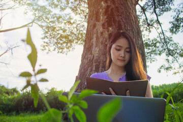 Front view of young girl sitting below big tree in park, she enjoying with reading book and using laptop, sunset light. Active on holiday concept.