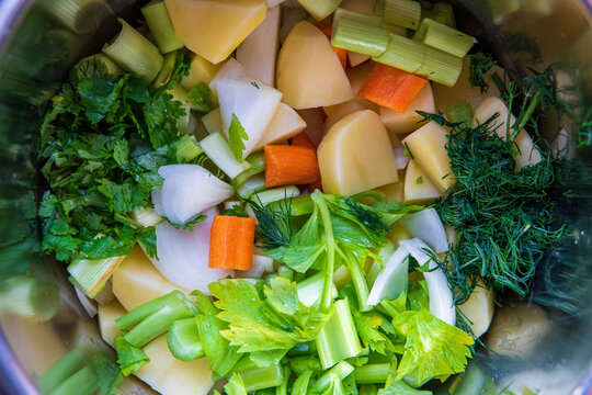 Fresh uncooked ingredients of vegetables and root veggies in a pot for cooking vegetables soup / stock. Potatoes, onions carrots, celery, leek, coriander, parsley and dill. top view