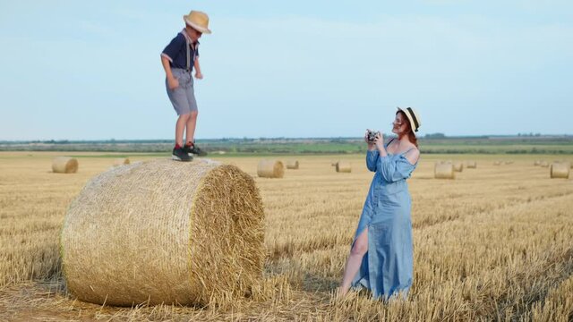 summer vacation, young happy mom takes pictures on retro camera of her cheerful son on haystack in field after harvesting against sky