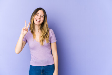 Young caucasian woman isolated on purple background showing victory sign and smiling broadly.