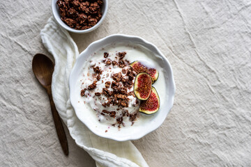 Granola mit frischen Feigen und Joghurt in einer weißen Schüssel. Beiges Leinen Tischtuch, Draufsicht, Stoffserviette, Holzlöffel.