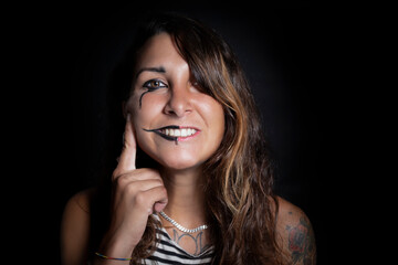 Portrait of a brown girl, tattooed, with face painted, smiling at the camera, with a black background.