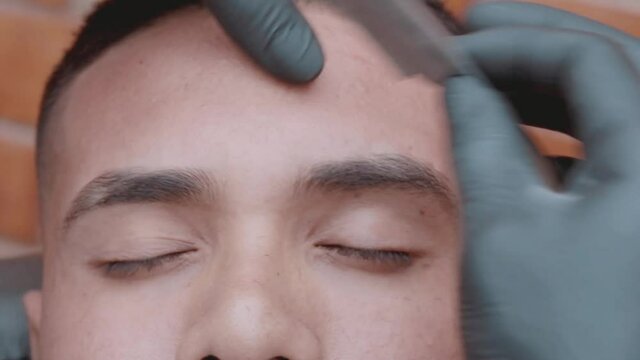 Barbero usando navaja y cepillo de peluquer&iacute;a para afeitar cejas de modelo masculino en peluquer&iacute;a 