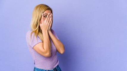 Young blonde woman isolated on purple background blink through fingers frightened and nervous.