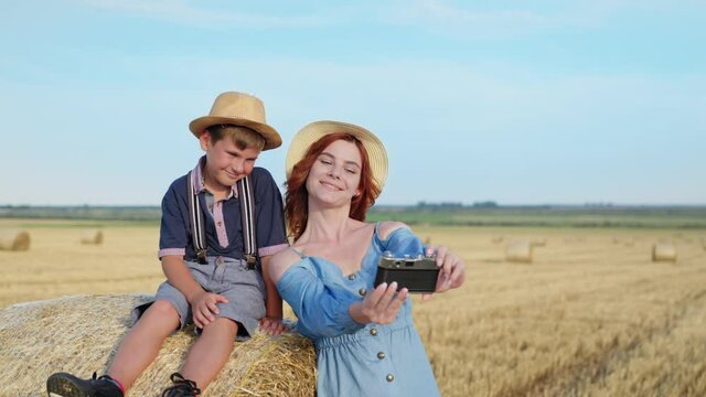 family photo, cute female parent and cute boy wearing straw hats have fun taking pictures with retro camera on haystack while relaxing in field after harvest