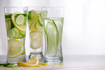 Three glasses of cucumber and lemon refreshing drink decorated with sliced lemon and cucumber on marble table with white background.