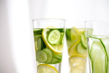 Closeup of three glasses of cucumber and lemon refreshing drink decorated with sliced lemon and cucumber on marble table with white background.