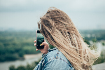 Young girl with long blond hair from behind. The girl looks into the phone camera. The girl shoots the landscape on the phone.
