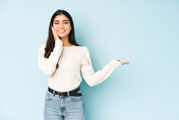 Young indian woman isolated on blue background holds copy space on a palm, keep hand over cheek. Amazed and delighted.