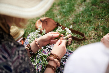 Close-up picture of woman, wearing colorful bracelets, making flower camomile wreath on green field on sunny summer day. Eco tourism in rural countryside. Young festival handmade activity.