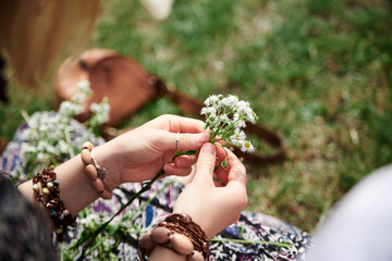 Close-up picture of woman, wearing colorful bracelets, making flower camomile wreath on green field on sunny summer day. Eco tourism in rural countryside. Young festival handmade activity.