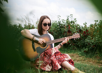 Young brunette hippie woman, wearing boho style clothes, sitting on green grass, holding guitar. Indie musician relaxing on green currant field on sunny summer day. Eco tourism concept.