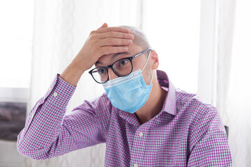 Mature man wearing protective mask, glasses and shirt checking his temperature with his hand on his forehead at home. Coronavirus first symptoms detection concept.