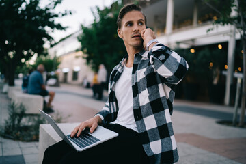 Serious caucasian male in casual wear sitting with laptop computer making mobile phone looking away, pensive skilled man digital nomad working remotely outdoors talking on smartphone on street
