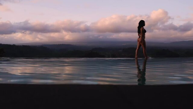 Playful mixed race woman toe walking on edge of infinity pool during sunset