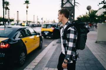 Rear view of caucasian male in trendy wear with backpack exploring city waiting for cab for transportation, 20s hipster guy standing on urban setting looking at cars and traffics in megalopolis