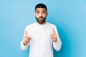 Young latin man against a blue  background isolated pointing upside with opened mouth.