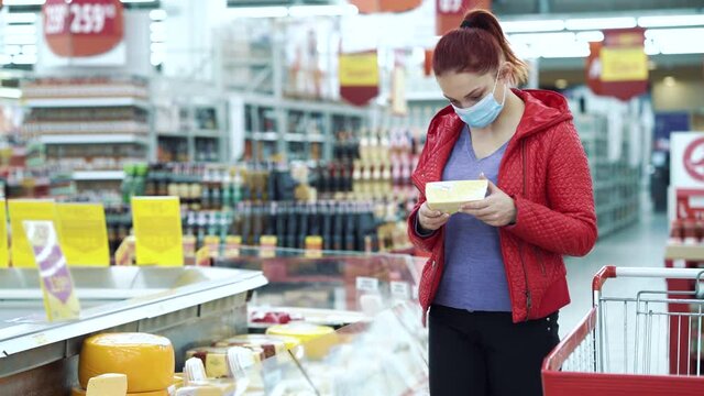 Red haired woman in red jacket wearing medical mask picking cheese from freezer in supermarket and putting it in trolley, blurred background. Customer buying food during pandemic. Concept of shopping