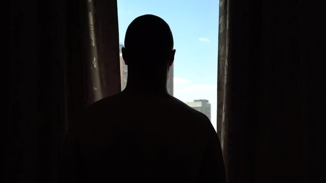 Man standing at home and tight-fitting curtains, silhouette against the window, social distance and self-isolation in quarantine isolation.