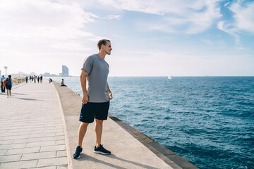 Smiling caucasian male runner recreating near water on quay during sunny morning feeling vitality and wellness, positive muscular handsome sportsman looking at river spending break on workout