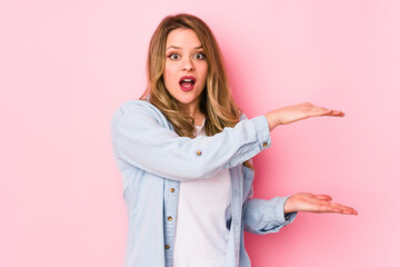 Young caucasian woman isolated on pink background shocked and amazed holding a copy space between hands.