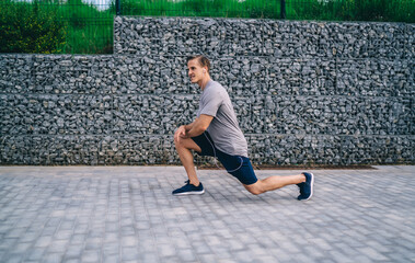 Strong male athlete doing squats stretching legs and lower body muscles on street during workout, ?aucasian sportsman keeping fit training doing warming exercises for getting vitality energy