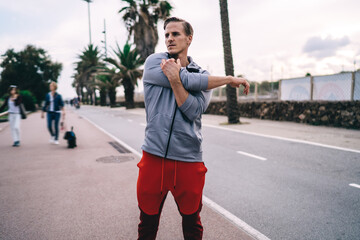 Fototapeta premium Muscular sportsman stretching body muscles during morning workout training at city street, Caucasian pumped up guy concentrated on practice warm up during intensive crossfit at urban setting