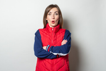 Young woman with a surprised face crossing her arms on her chest in a red vest on a light background