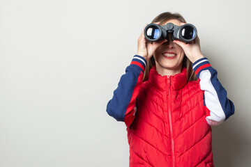 Young woman in a red vest looks through binoculars on a light background