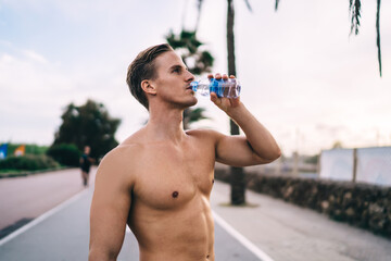 Handsome Caucasian male jogger with perfect body drinking water after workout outdoors standing on...