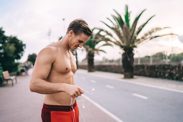 Caucasian male jogger with perfect shirtless press holding electronic earphones and smiling, successful sportsman with headphones enjoying leisure time for physical recreation at urban street