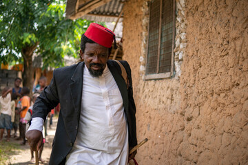 An African Older Man in Red Muslim Taqiyyah Fez Hat And Blazer on white Dress Moving a stick for lame people Near the Basic Hut with Thatched roof in Small Remote Village in Tanzania, Pemba island