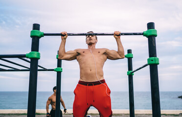 Strong athlete in earphones have morning workout at sports playground with bars for training...