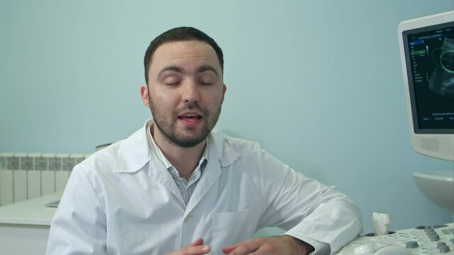 Young Male Doctor Talking To The Camera Sitting Next To Ultrasound Scanning Machine