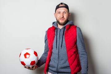 Young man in a cap, hoodie and vest holds a ball in his hand on a light background