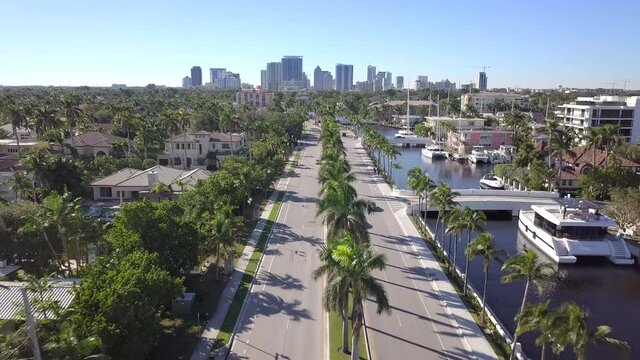 aerial drone following luxurious GT car on road with palm trees in Luxury neighborhood, buildings and yacht's in the background